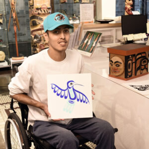 Pacific Northwest Coast Native Artist Adonis David sitting his his wheel chair showing one of his hummingbird acrylic paintings